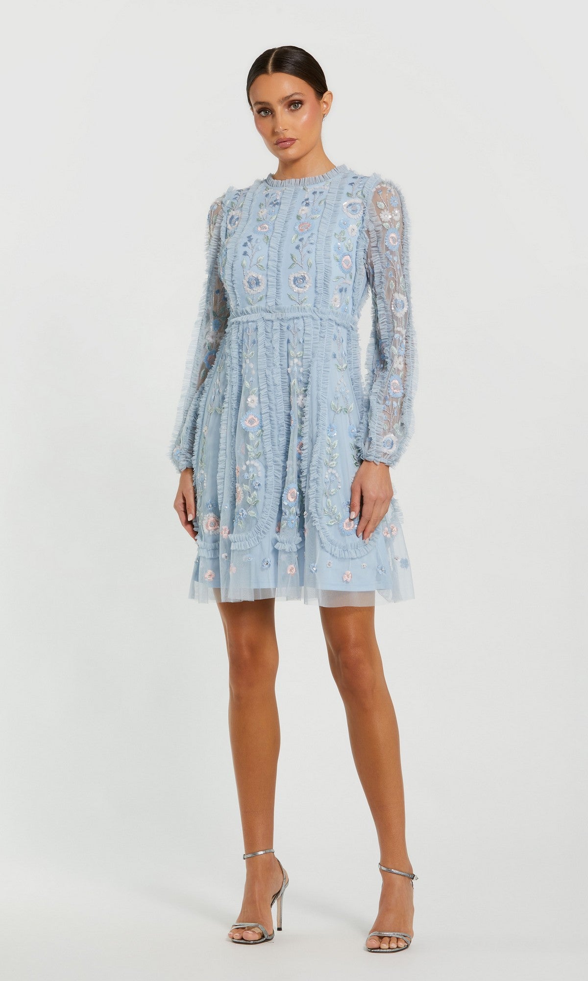 A woman stands against a plain white background wearing the powder blue Mac Duggal 9365 short wedding guest dress with sheer embroidered sleeves and silver high-heeled sandals.